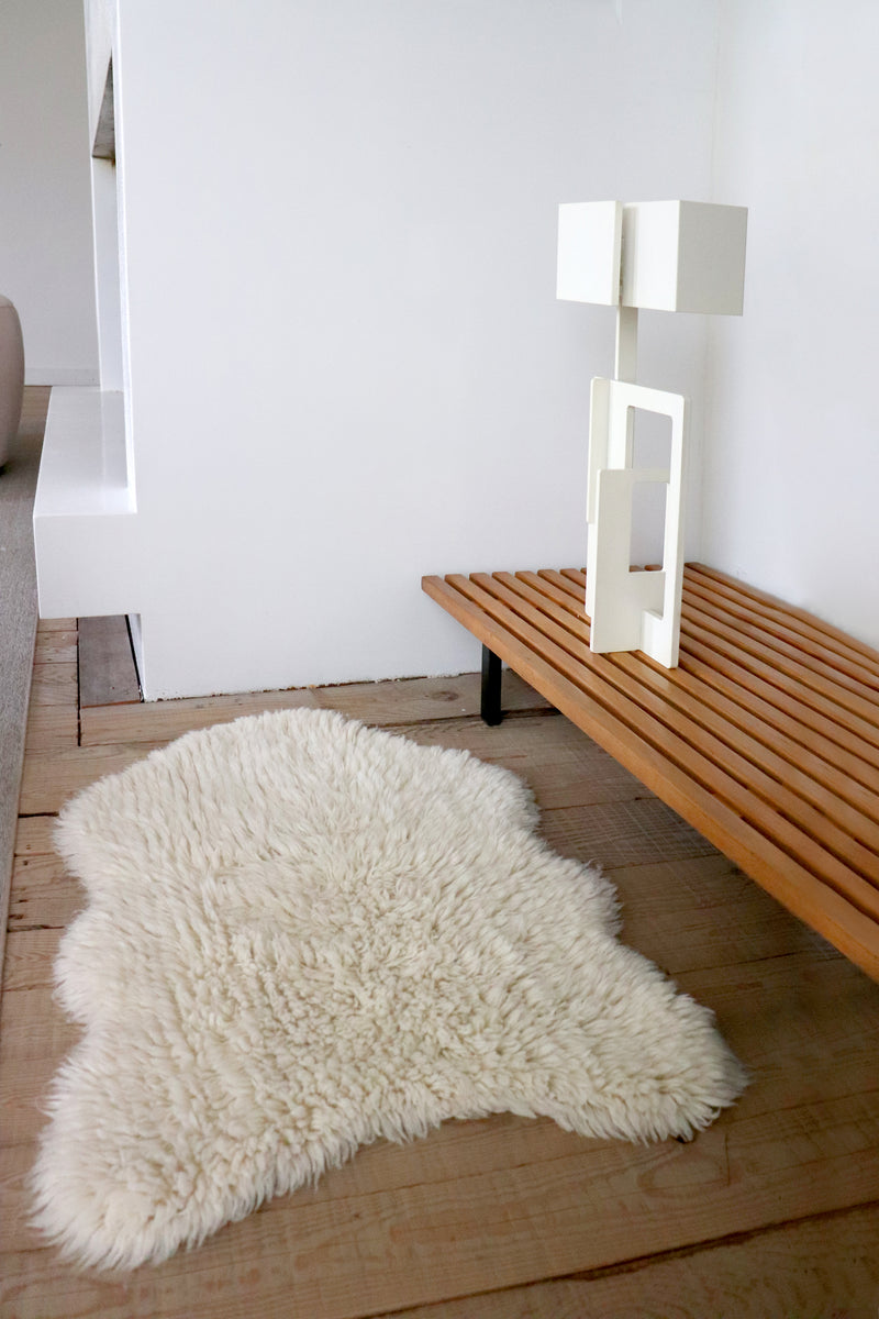 White fluffy sheep rug on a wooden floor next to a wooden bench and white wall.