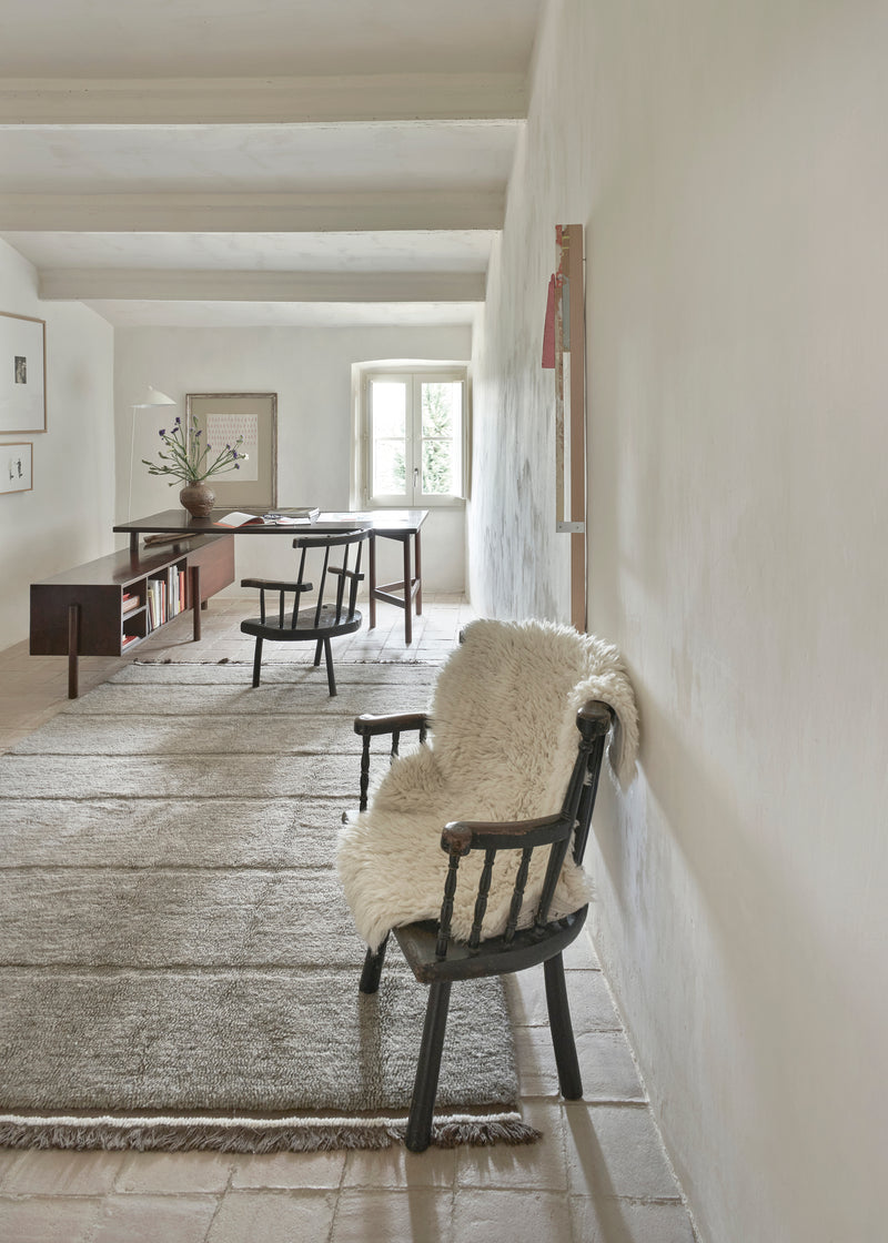 Narrow hallway with a chair draped with a undyed sheep rug, leading to a room with a desk and window.