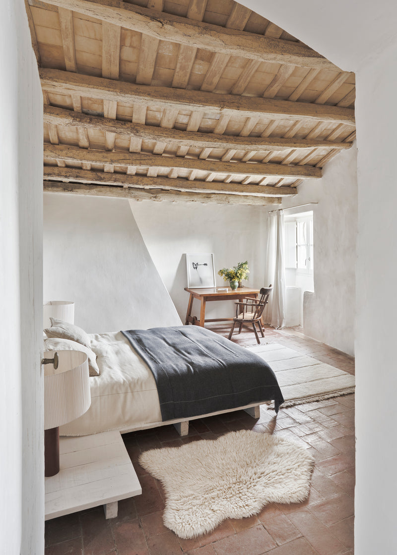 Cozy bedroom with wooden beams on the ceiling, bed with white bedding, sheepskin rug on the floor, and a small table with a plant.