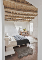 Cozy bedroom with wooden beams on the ceiling, bed with gray blanket, and small table with plants.