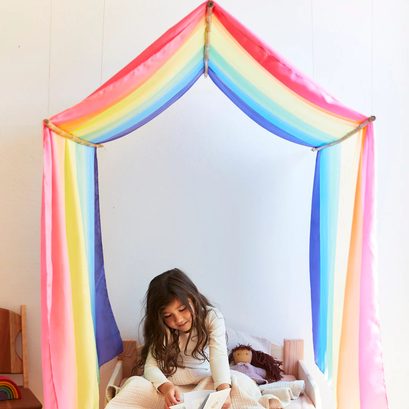 Child sitting inside a colorful rainbow tent in a room.